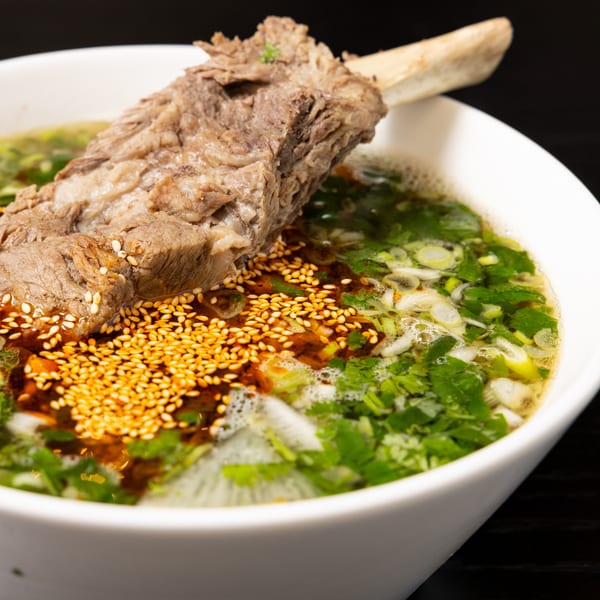 Halal beef rib soup with green onions, cilantro, chili oil, and sesame seeds in a white bowl.
