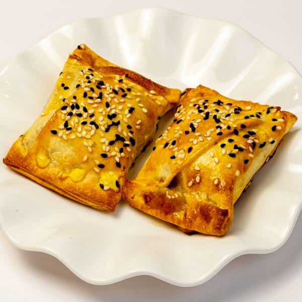 Two golden-brown Kusan Collective pastries, topped with black and white sesame seeds, on a white ruffled plate.