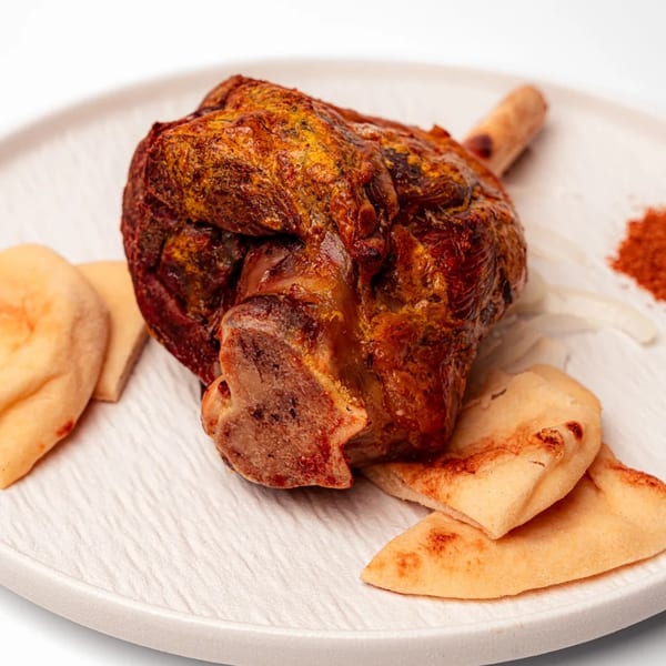 A roasted lamb shank with golden-brown skin, served with flatbread and spices on a white plate.