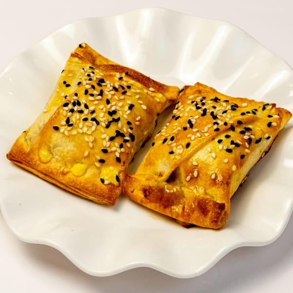 Two golden-brown, sesame-topped pastries, possibly Kusan Collective's halal samosas, on a white ruffled plate.